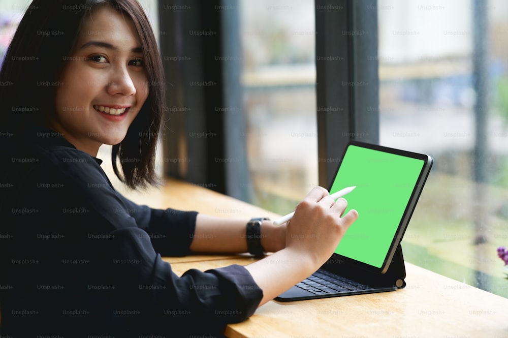 Young woman using stylus pen pointing on black tablet with green screen while sitting in office.