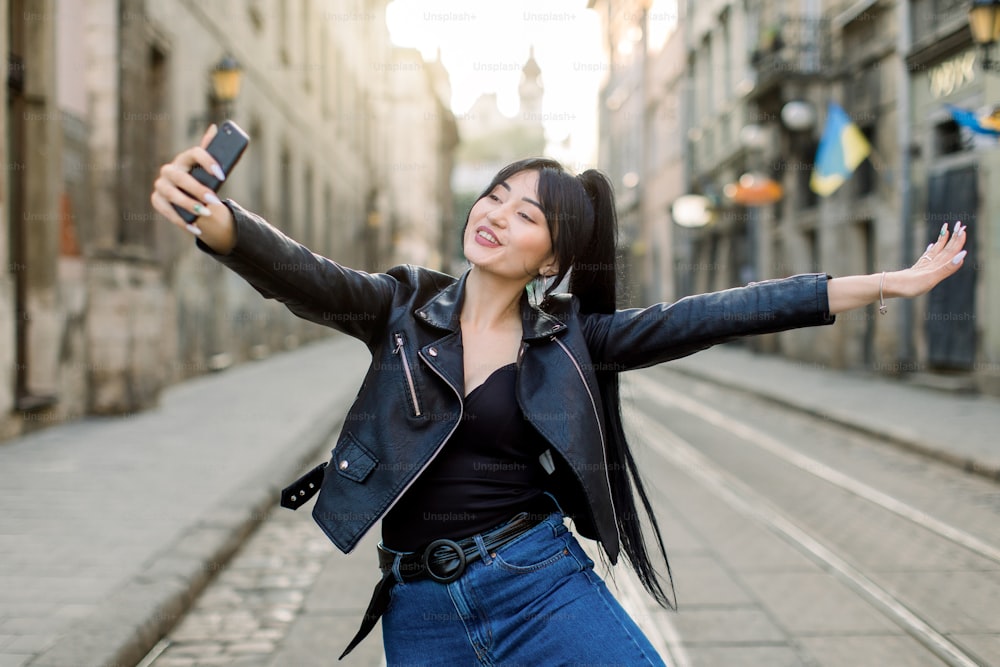 Happy female mixed raced tourist taking selfie on city street, smiling and having fun, enjoying her walk. Young Asian lady in fashionable clothes making selfies photo on the phone.