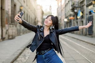 Happy female mixed raced tourist taking selfie on city street, smiling and having fun, enjoying her walk. Young Asian lady in fashionable clothes making selfies photo on the phone.