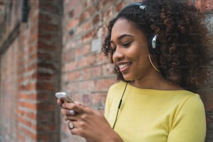 Portrait of young Afro american woman using mobile phone and listening to music with headphones in the street. Outdoors.