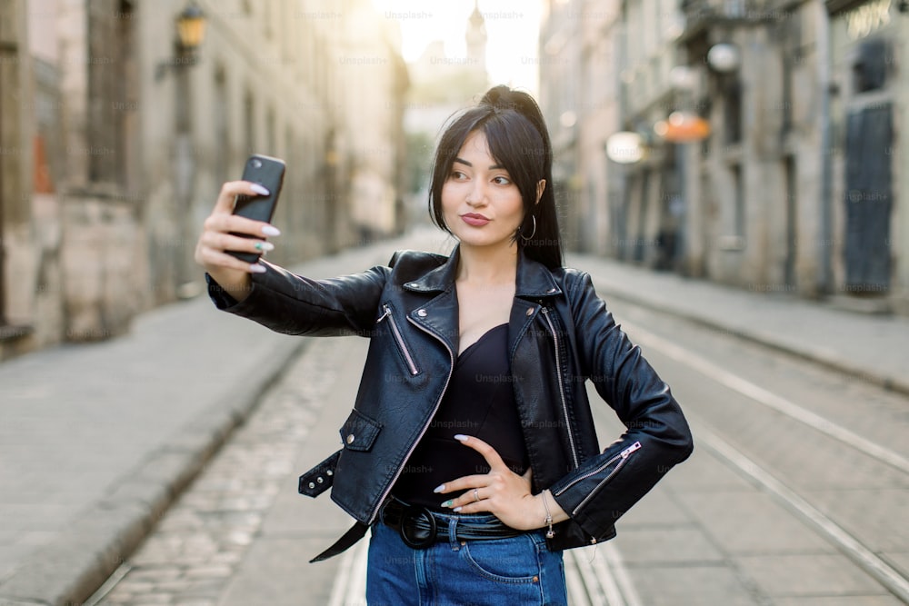 ジーンズと黒い革のジャケットを着た若いセクシーで魅力的なアジアの女性は、道路に立って、街の屋外でポーズをとって、彼女のsmarphoneで自撮り写真を作ります。人、ライフスタイル、都市のコンセプト。