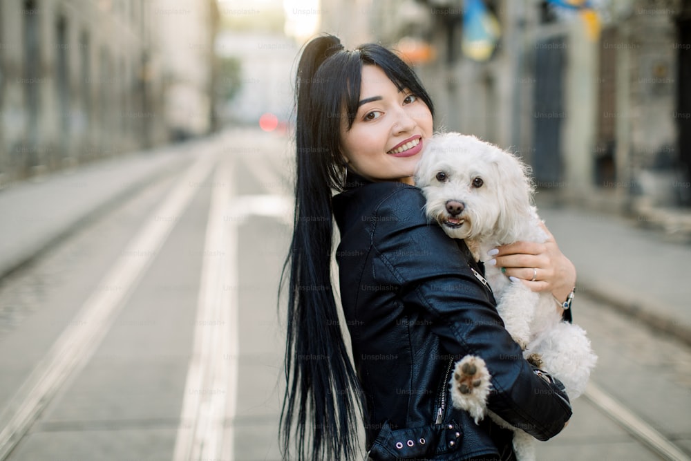 Toma horizontal al aire libre de una mujer asiática con peinado de pony oscuro, con chaqueta de cuero, sonriendo a la cámara mientras camina por la calle de la ciudad vieja con su lindo perrito blanco.