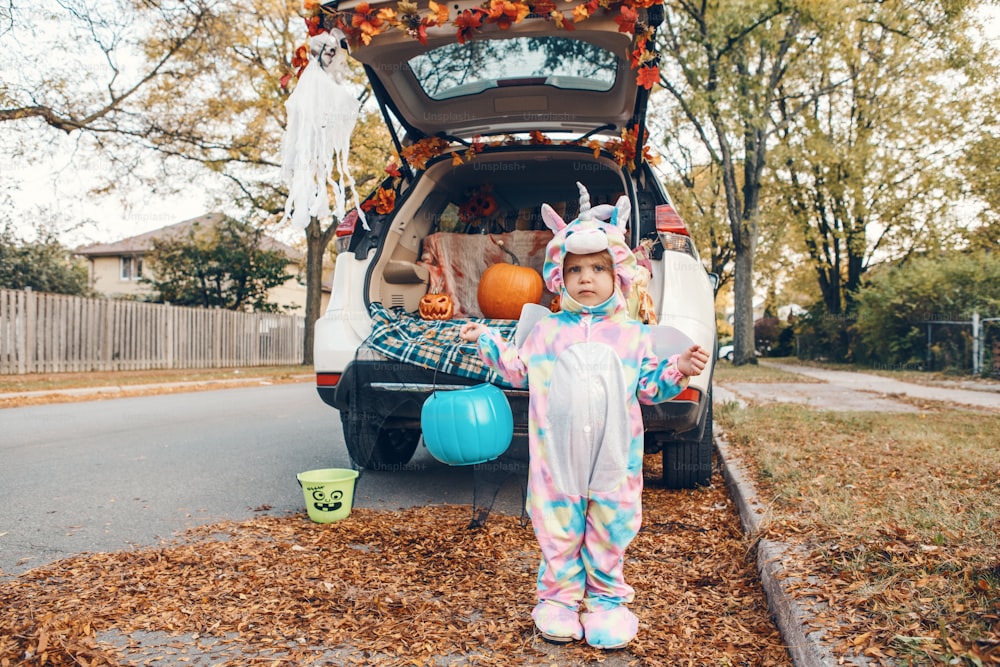 트릭 또는 트렁크. 자동차 트렁크에서 할로윈을 축하하는 유니콘 의상을 입은 슬픈 화난 아기. 10월 휴일 야외를 준비하는 귀여운 유아. 사회적 거리와 안전한 대안 축하.