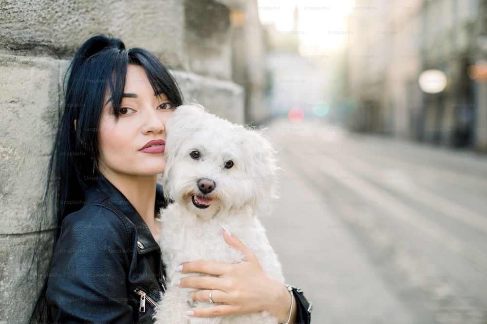 Pretty Asian woman model wearing stylish black leather jacket, looking at camera, while walking in old city with her cute little white dog. Sunny morning. Close up. City lifestyle. Copy space