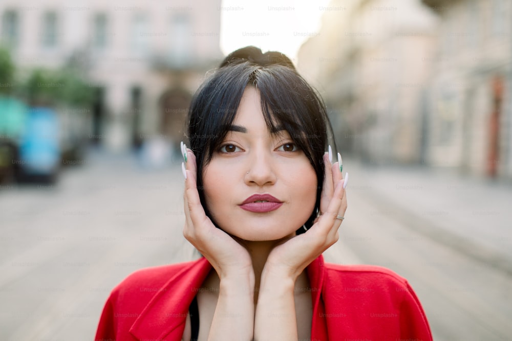 Close-up face outdoor portrait of pretty brunette Asian female model with  black hair and fashion make up, leaning face on her palms, posing at camera  at old city street background. photo –