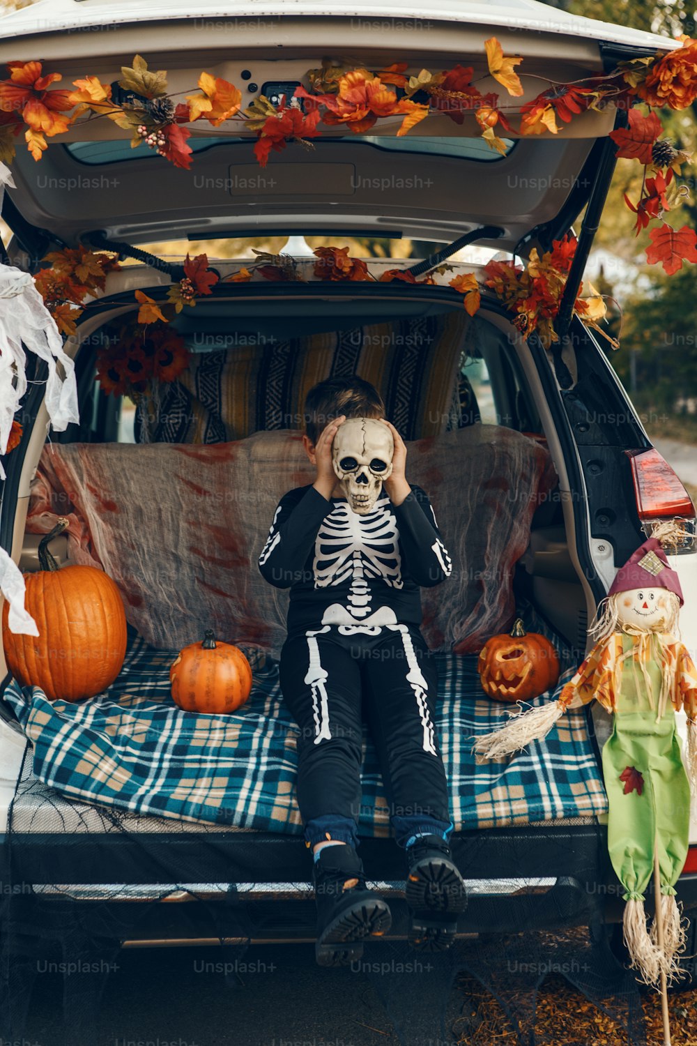 Trick or trunk. Boy child with human skull celebrating Halloween in decorated trunk of car. Happy kid preparing for October holiday outdoor. Social distance and safe alternative celebration.