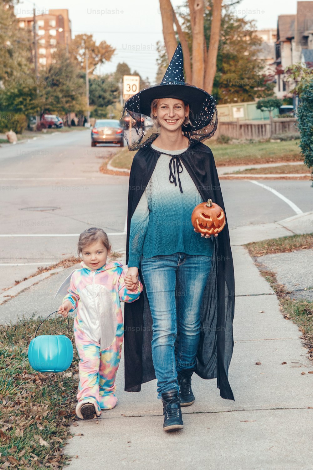 Trick or treat. Mother with baby toddler girl going to trick or treat on Halloween holiday. Mom with kid child in party costumes with basket going to neighbourhood houses for candies, treats.