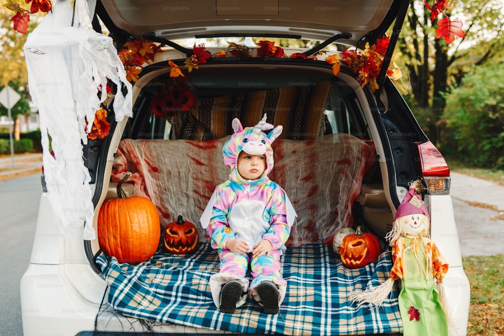 Trick or trunk. Sad upset baby in unicorn costume celebrating Halloween in trunk of car. Cute toddler celebrating October holiday outdoor. Social distance and safe alternative celebration.