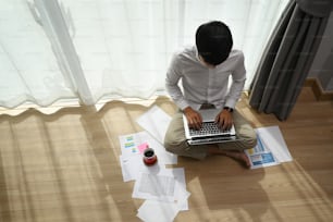Overhead view of young man working typing on laptop while sitting on the floor at modern home.