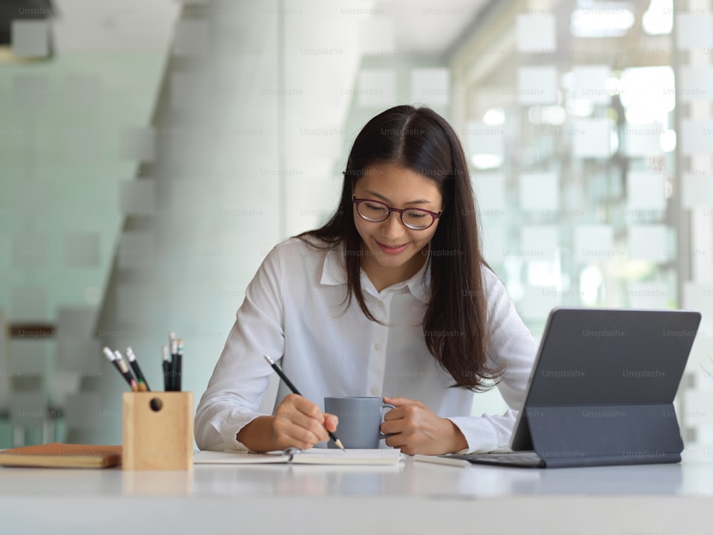 Professional businesswoman writing on notebook while holding a coffee cup in workspace