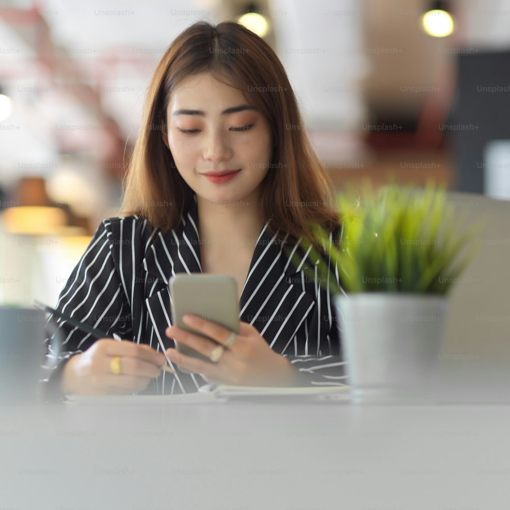Portrait of female office worker using smartphone while sitting at worktable in office room