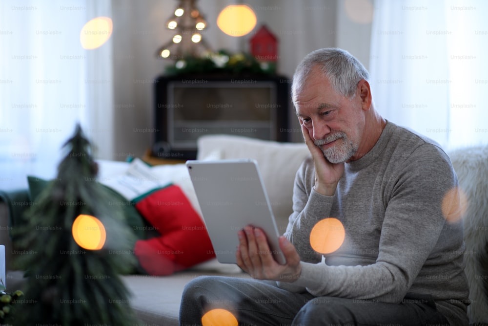 Porträt eines traurigen und einsamen älteren Mannes mit Tablet, der zu Weihnachten drinnen am Tisch sitzt, Einsamkeitskonzept.