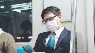 Reisende tragen Gesichtsmaske, während sie ihr Mobiltelefon im öffentlichen Zug benutzen. Coronavirus-Krankheit oder COVID-19-Pandemie-Ausbruch und städtisches Stadt-Lifestyle-Problem im Berufsverkehrs-Pendlerkonzept.
