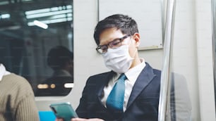 Traveler wearing face mask while using mobile phone on public train . Coronavirus disease or COVID 19 pandemic outbreak and urban city lifestyle problem in rush hour commuting concept .