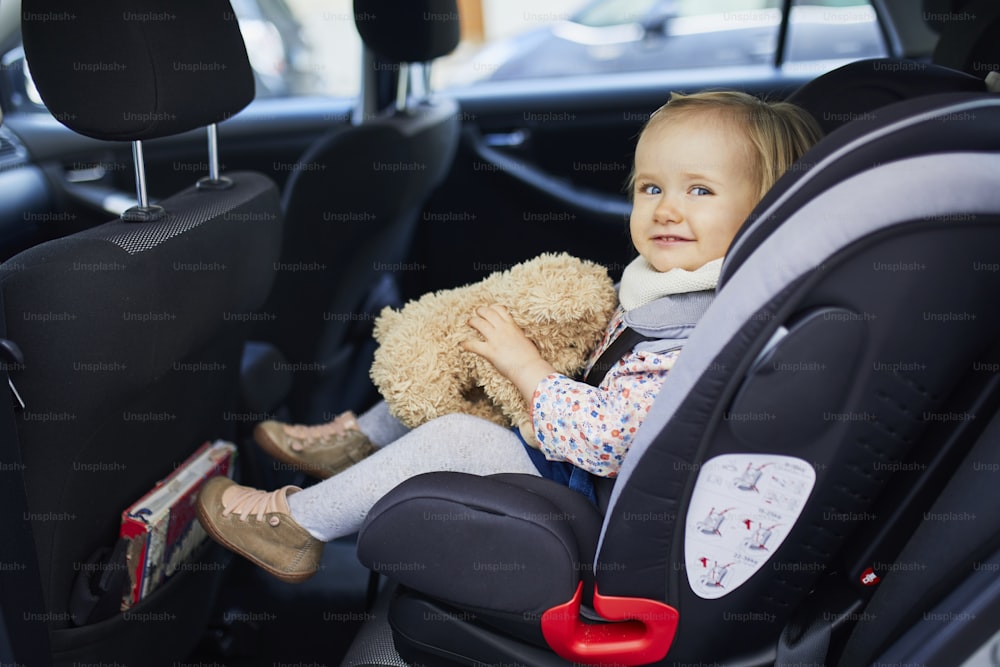 Adorable toddler girl in modern car seat with her favorite stuffed toy. Little kid traveling by car. Child safety on the road. Trip with a baby
