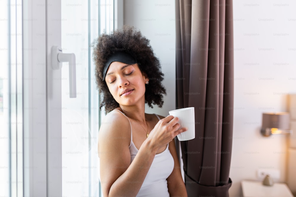 Beautiful young woman drinking coffee and looking through window while sitting at windowsill at home having a perfect cozy morning.