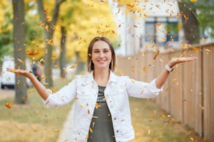 Happy middle age Caucasian woman throwing autumn fall yellow leaves outdoor. Young woman with short haircut in casual clothes enjoying life in park outside. Little memorable moments of life.