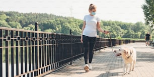 Caucasian woman with medical mask on face is walking with her dog near a lake in park during a summer hot day