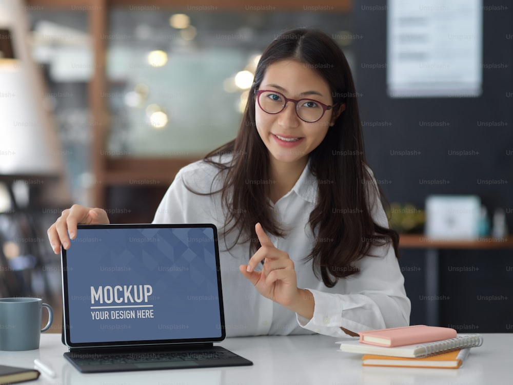 Young pretty businesswoman pointing at blank screen mock up tablet in the office room, include clipping