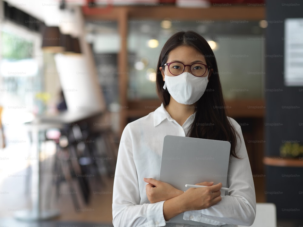 Young businesswoman standing in her modern office room while wearing a mask