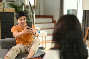 Young man using smart phone taking photo of his girlfriend on couch in living room.