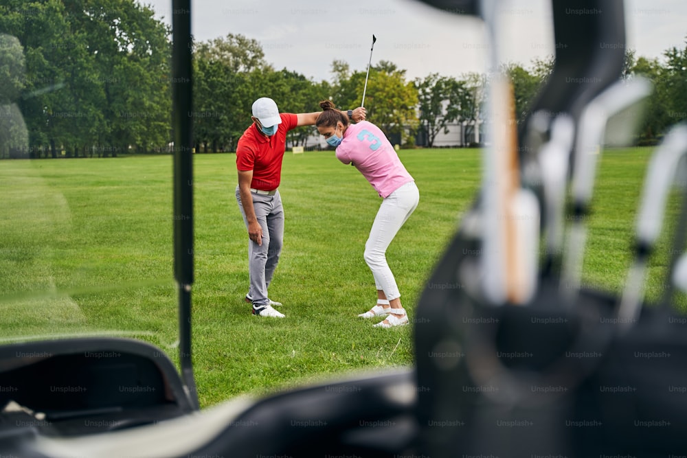 Concentrated beginner golfer attempting to hit the ball with an iron club helped by her trainer