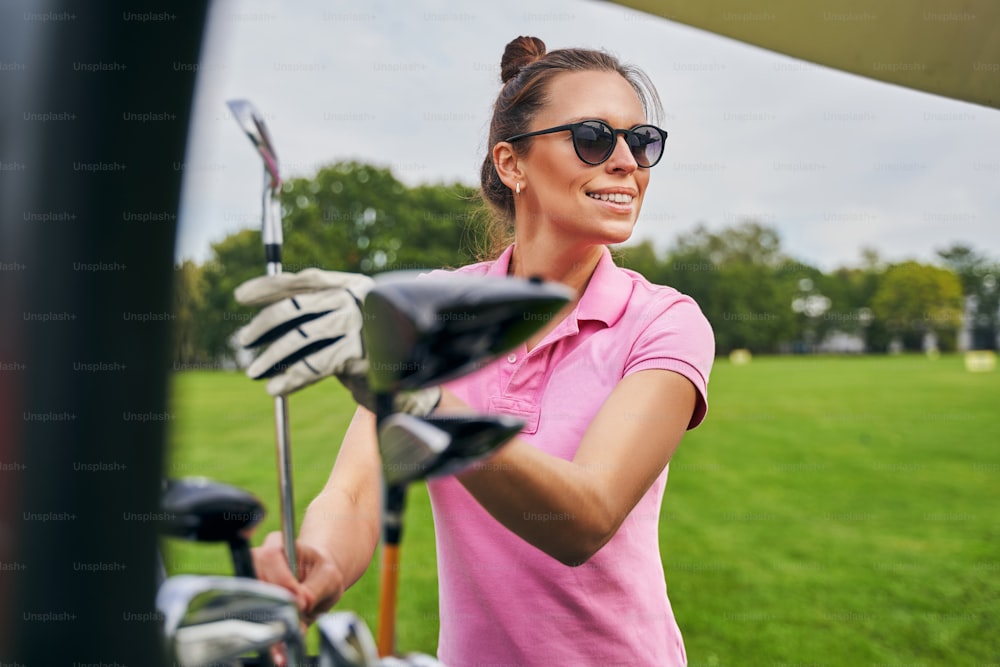 Waist-up portrait of a joyous woman in a polo shirt holding a wedge club with both hands
