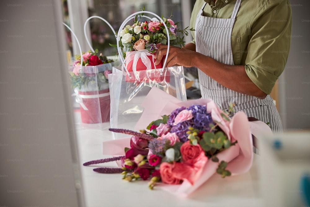 Cropped photo of a flower shop salesperson putting a pot of blooming flowers in a plastic bag near a bouquet