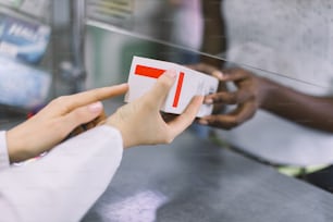 Medicina, farmacia, salud y concepto de personas. Imagen recortada de cerca de las manos de una farmacéutica que da una caja blanca con un medicamento a una clienta africana en la farmacia.