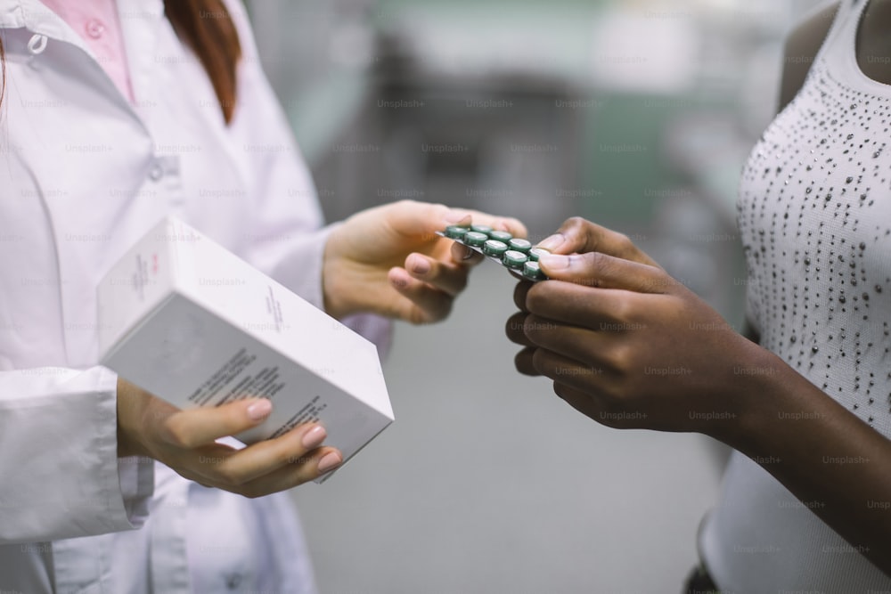 여성 약사가 마약이 담긴 상자를 들고 흑인 여성에게 알약으로 물집을 주고 현대 커뮤니티 약국에서 약을 조제하는 손의 자른 이미지.