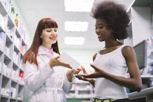 Pretty young Caucasian red haired pharmacist advising her young African female client at modern pharmacy how to take medicine. Medicine, pharmaceutical care concept.