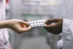 Nahaufnahme zugeschnittenes Bild der Hände einer afrikanischen Apothekerin, die der kaukasischen Patientin die Blisterpackung mit weißen Pillen gibt. Medikamentenabgabe, pharmazeutische Versorgung.