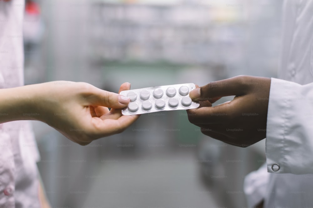 Close up cropped image of hands of female African pharmacist, giving the blister with white pills to female Caucasian patient. Drug dispensing, pharmaceutical care.