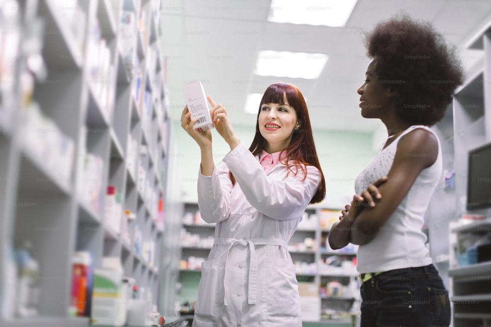 Young Caucasian cheerful woman pharmacist is recommending medicine for young black woman client in modern drugstore. Female pharmacist consulting a customer in pharmacy, showing the drug.