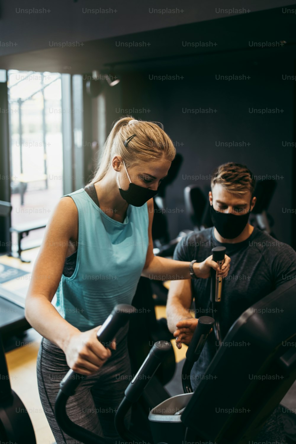 Young fit and attractive woman at body workout in modern gym together with her personal fitness instructor or coach. They keeping distance and wearing protective face masks. Coronavirus world pandemic and sport theme.