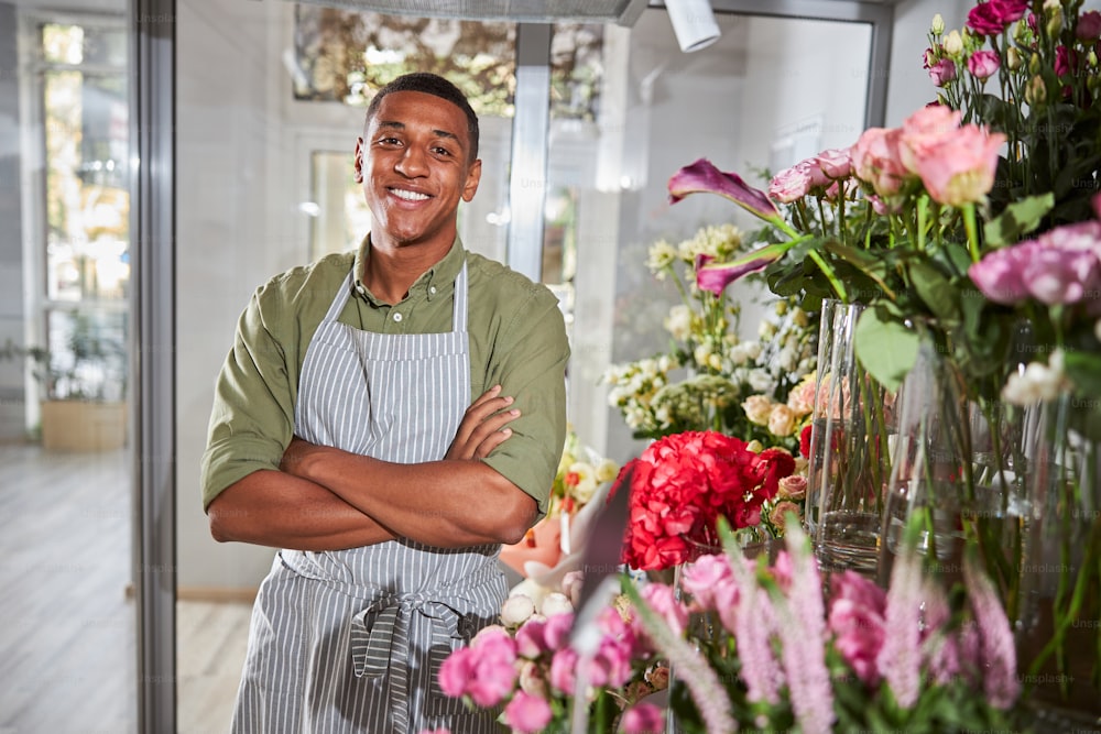 Optimistic florist standing near a big flower display with his arms crossed below his chest