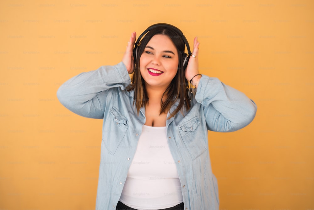 Portrait of young plus size woman listening to music with headphones outdoors against yellow background.