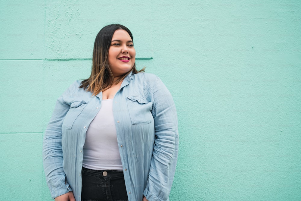 Close-up of young plus size woman smiling while standing against light blue wall outdoors. Urban concept.