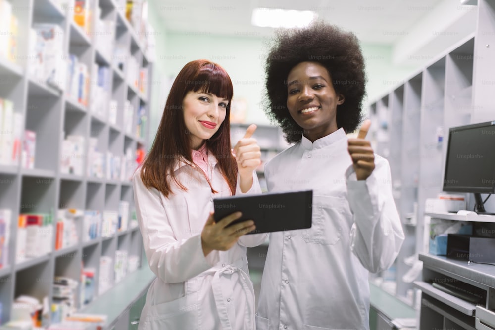 Zwei selbstbewusste multiethnische Apothekerinnen in der modernen Pharmazie. Afroamerikanische und kaukasische Chemikerinnen, die Notizen auf dem Tablet machen und Daumen hoch während der Inventur in der Apotheke zeigen.