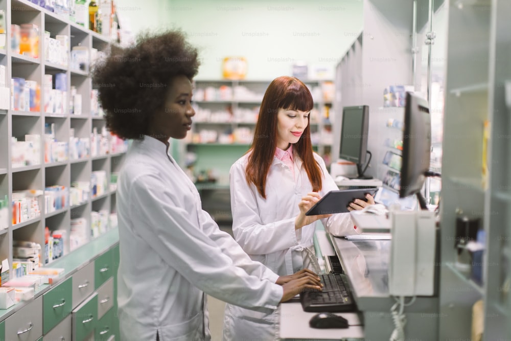 Ritratto di due sorridenti farmaciste multietniche amichevoli che lavorano su computer e tablet in farmacia moderna e fanno ordine per i medicinali nella società di distribuzione.