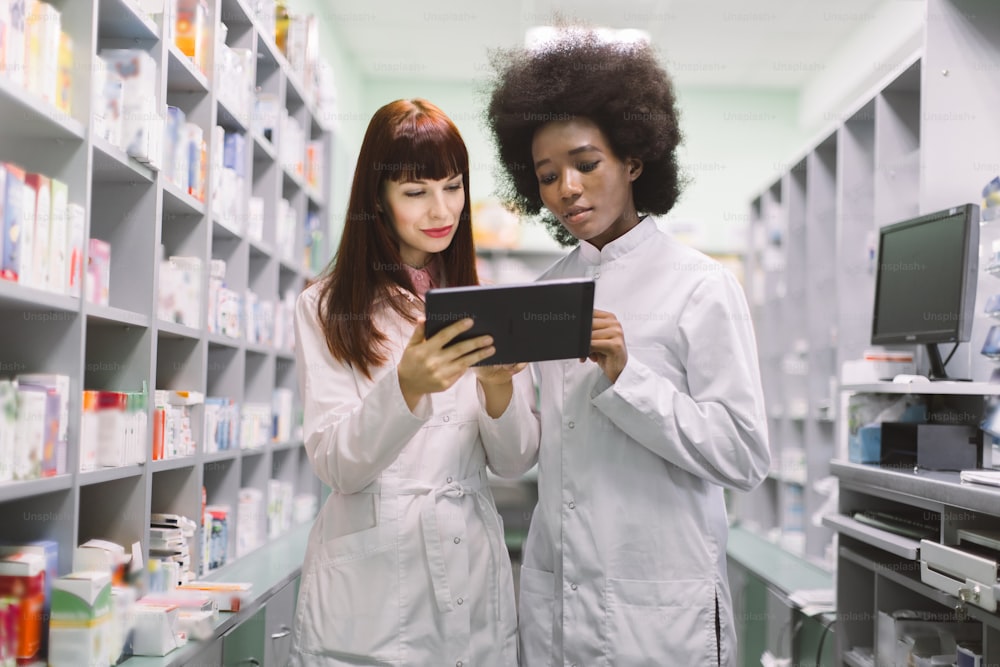 Due giovani farmaciste multirazziali sicure di sé, africane e caucasiche, in farmacia moderna, che guardano lo schermo del tablet pc, mentre controllano le medicine o fanno un ordine.