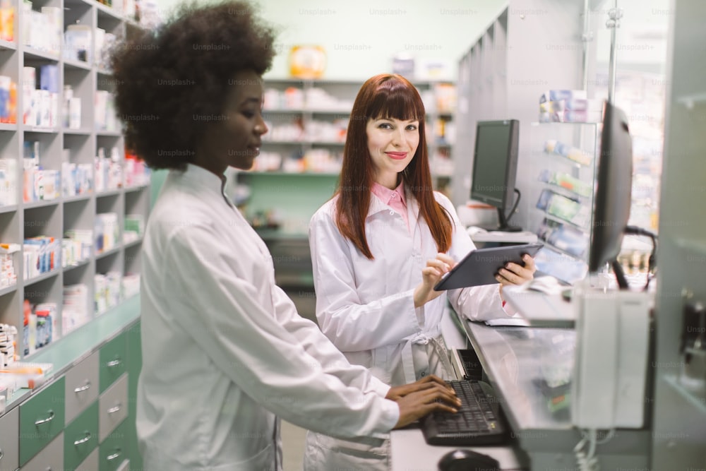 Dos jóvenes y alegres farmacéuticas que trabajan juntas en la farmacia. Mujer africana escribiendo en la computadora mientras su colega caucásico usa la tableta. Enfócate en la mujer caucásica.