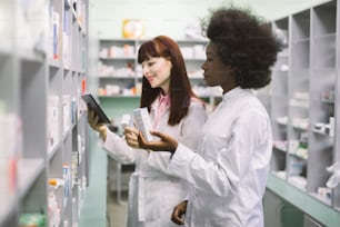 Medicine, pharmaceutics, health care and people concept. Two young multiethnic female pharmacist, African and Caucasian, taking medications from the shelf, using tablet in pharmacy.