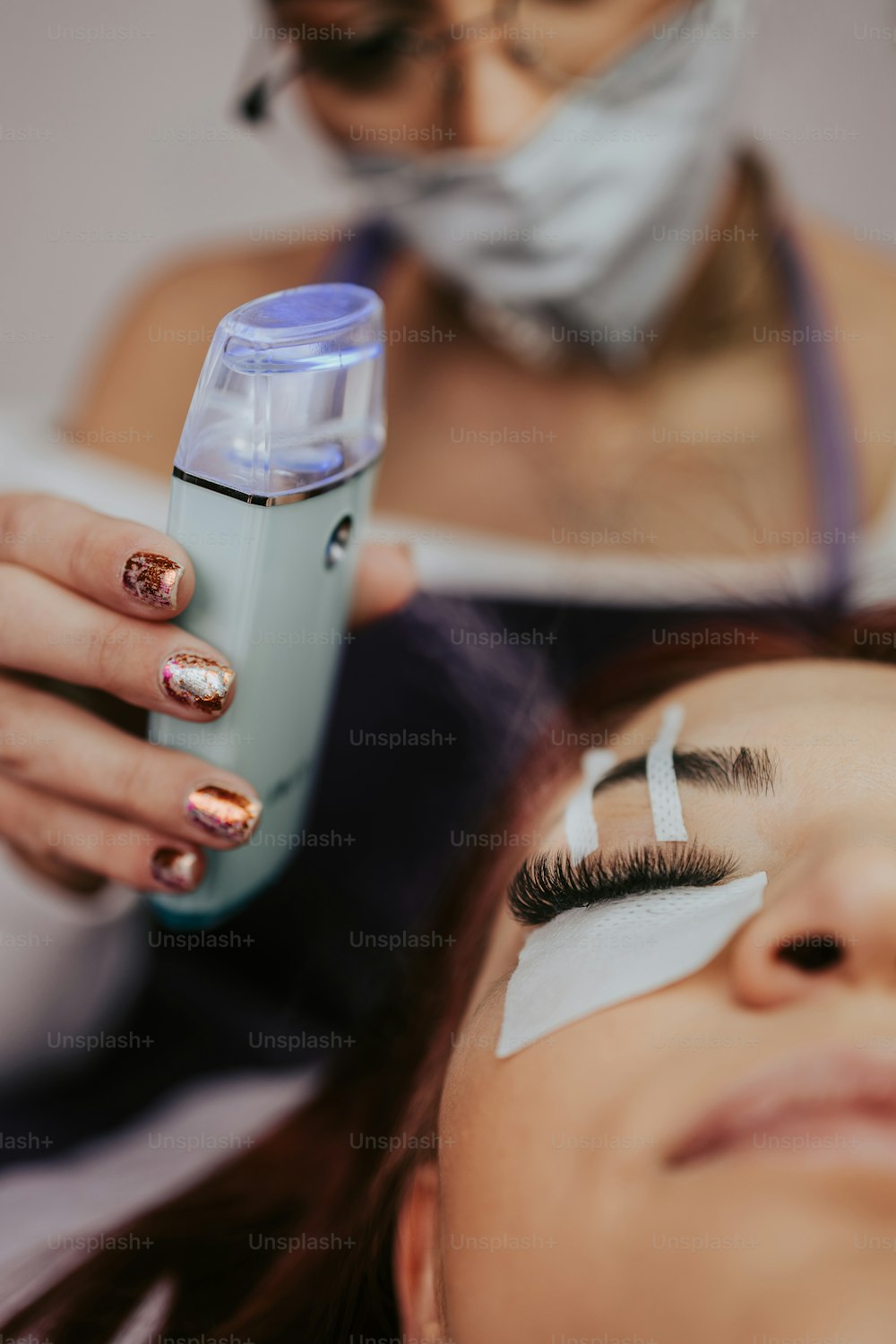 Beautiful young woman at eyelash extension procedure. Cosmetics and body care close up shot. Female beautician wearing protective face mask. Coronavirus stay safe concept.