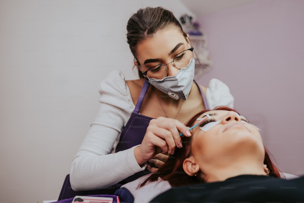 Beautiful young woman at eyelash extension procedure. Cosmetics and body care close up shot. Female beautician wearing protective face mask. Coronavirus stay safe concept.