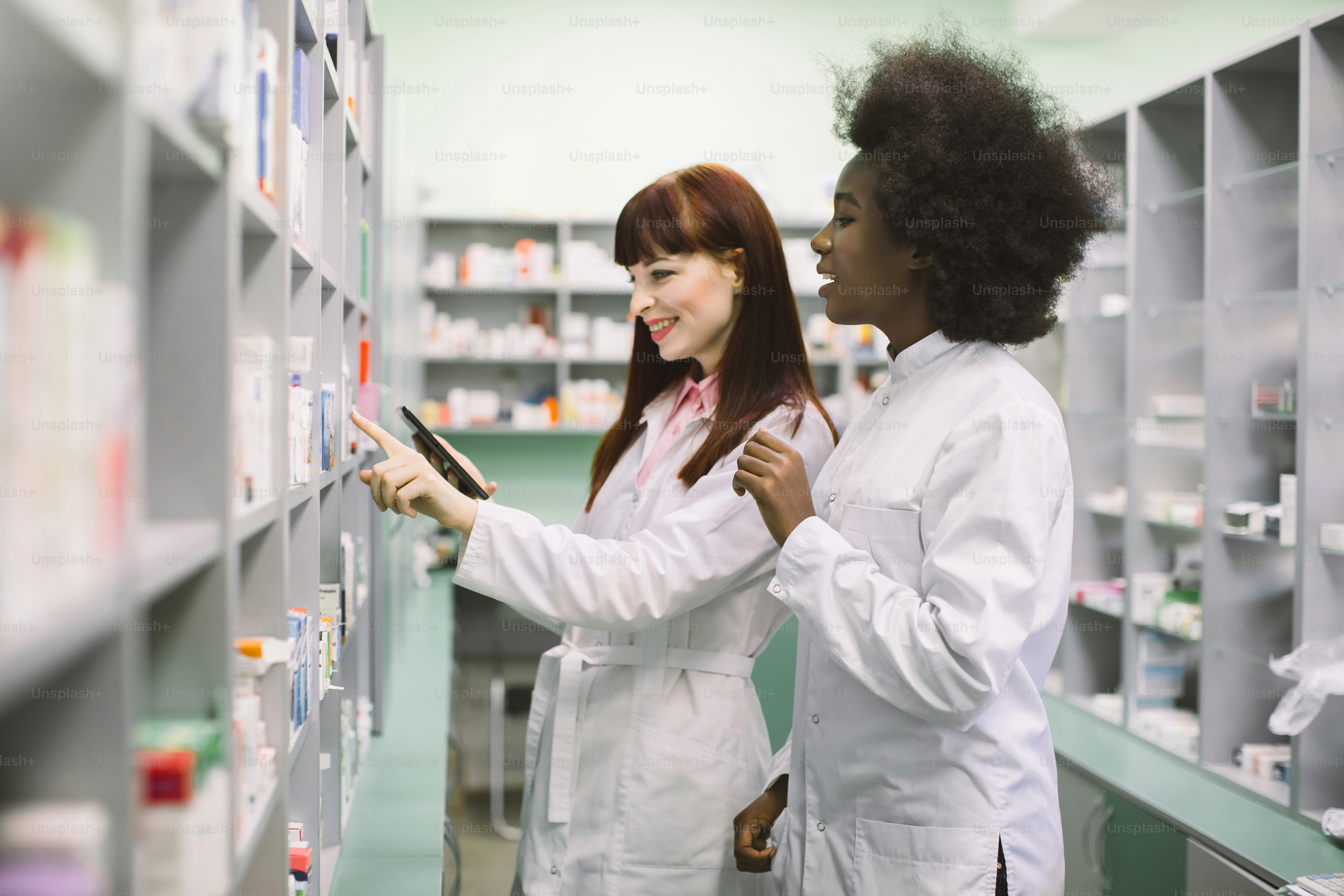 Zwei professionelle multiethnische Apothekerinnen, afrikanische und kaukasische Frauen, überprüfen den Medikamentenbestand mit Tabletten, während sie im Regal in einer modernen Drogerie stehen.