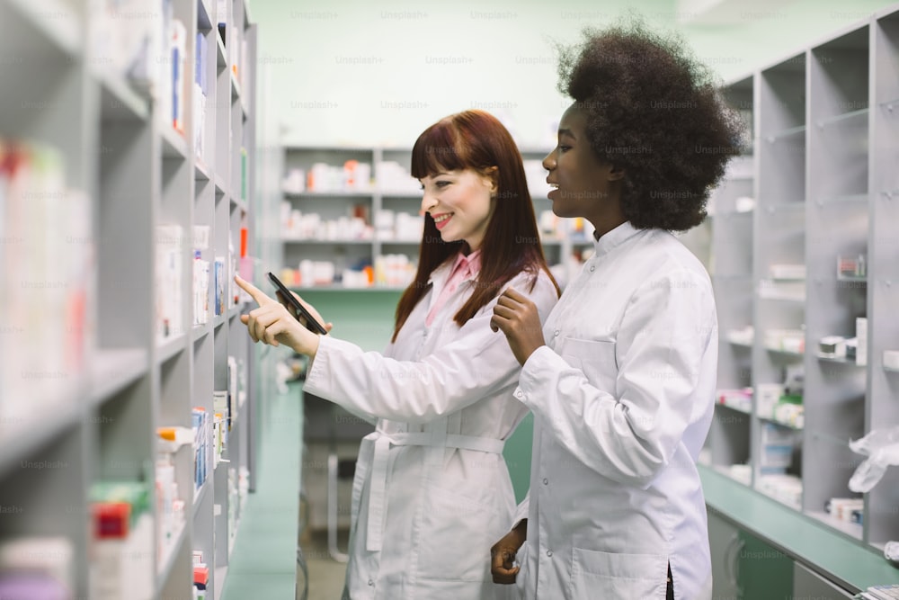 Two professional multiethnical pharmacists, African and Caucasian women, checking stock of medicines, using tablet, while standing at the shelve in modern drugstore.