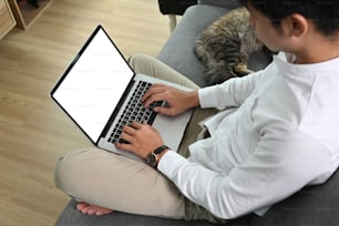 Foto de cabeza de un hombre usando una computadora portátil con pantalla blanca mientras está sentado en el sofá con su gato.