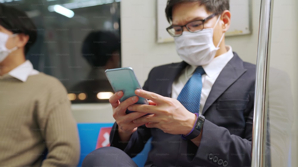 公共の電車で携帯電話を使用している間、フェイスマスクを着用している旅行者。コロナウイルス病またはCOVID19パンデミックの発生とラッシュアワーの通勤コンセプトにおける都市ライフスタイルの問題。