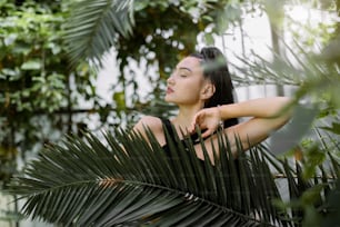 Beautiful mixed raced Asian woman model with dark long ponytail hair style, wearing black dress, posing in greenhouse with tropical plants, standing behind palm tree leaf. Tropical, summer concept